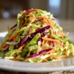 A mound of fresh, colorful cabbage salad featuring shredded green cabbage, purple cabbage, and carrots, served on a white plate.