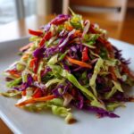 A vibrant mound of shredded cabbage salad featuring green and purple cabbage, carrots, and red peppers tossed in a light dressing.