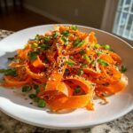 A close-up of a vibrant raw carrot salad made with shaved carrot ribbons, topped with sesame seeds and green onions.