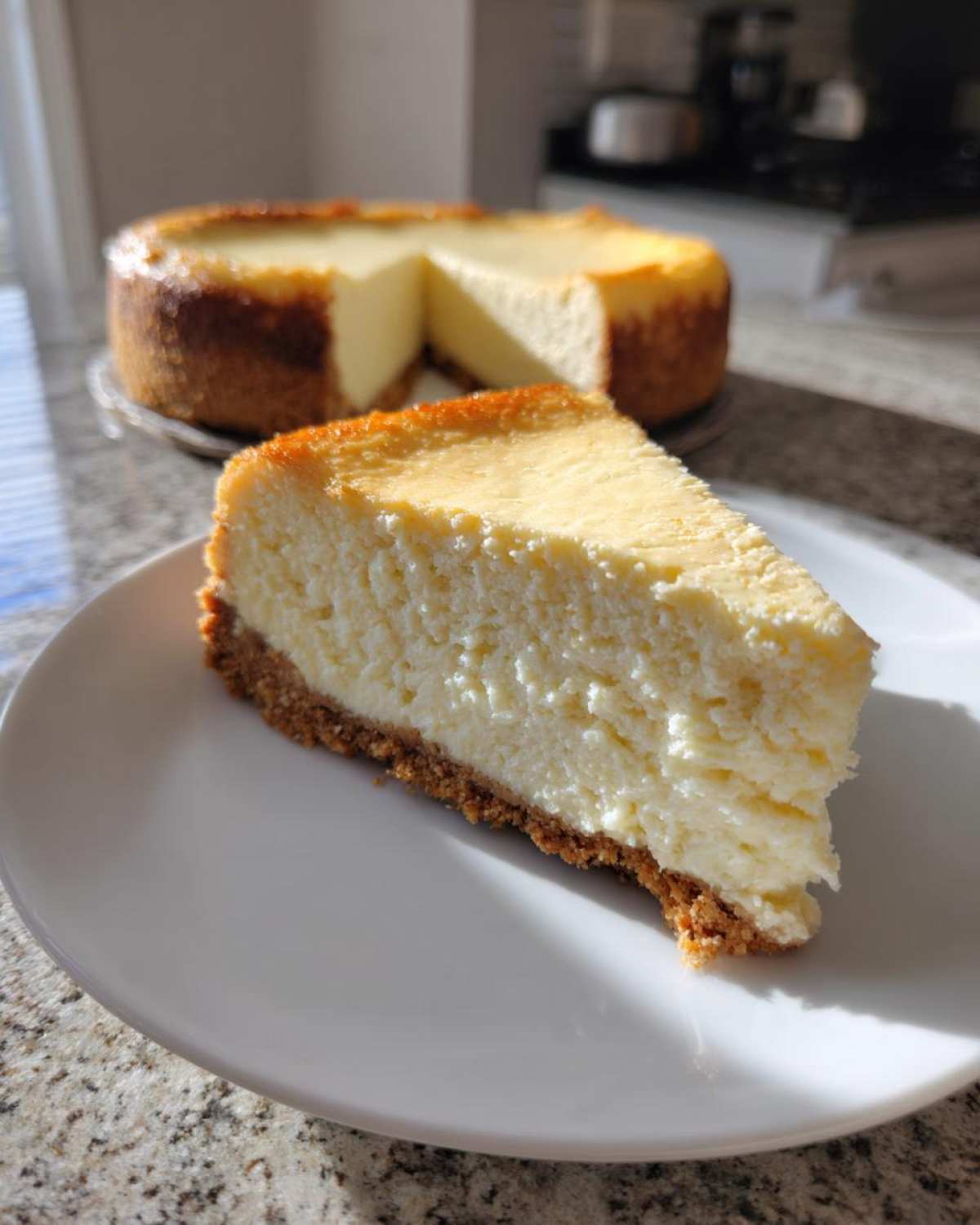 A close-up slice of creamy white chocolate cheesecake on a white plate, with the rest of the baked cheesecake in the background.