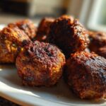 Close-up of several browned Air Fryer Italian Meatballs resting on a white plate, catching bright sunlight.