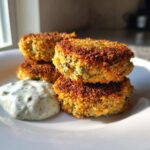 Four crispy, golden Air Fryer Salmon Patties stacked on a white plate next to a dollop of creamy dill sauce.