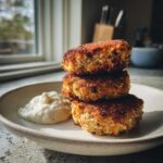 Three golden brown, crispy Air Fryer Salmon Patties stacked on a plate next to a dollop of white dipping sauce.