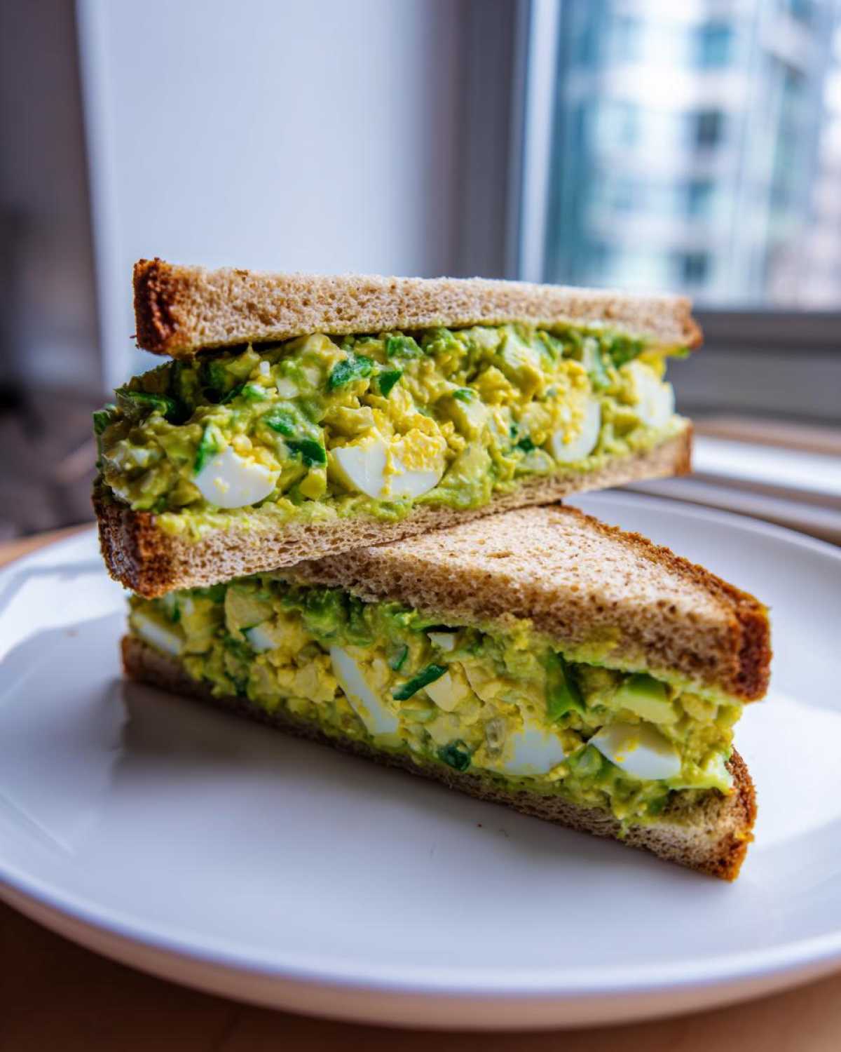 Two halves of an Avocado Egg Salad Sandwich stacked on a white plate, showing creamy green filling and egg chunks.