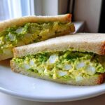 Two halves of an Ultimate Avocado Egg Salad Sandwich showing chunky egg and bright green avocado filling on white bread.