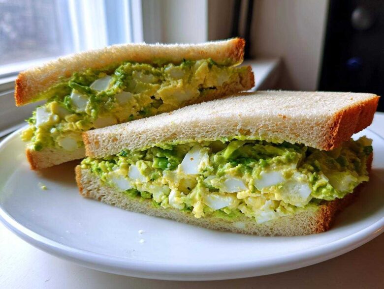 Two halves of an Ultimate Avocado Egg Salad Sandwich showing chunky egg and bright green avocado filling on white bread.