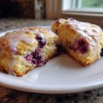 Two halves of a flaky Blueberry Lemon Scones, drizzled with a sweet white glaze, resting on a white plate.