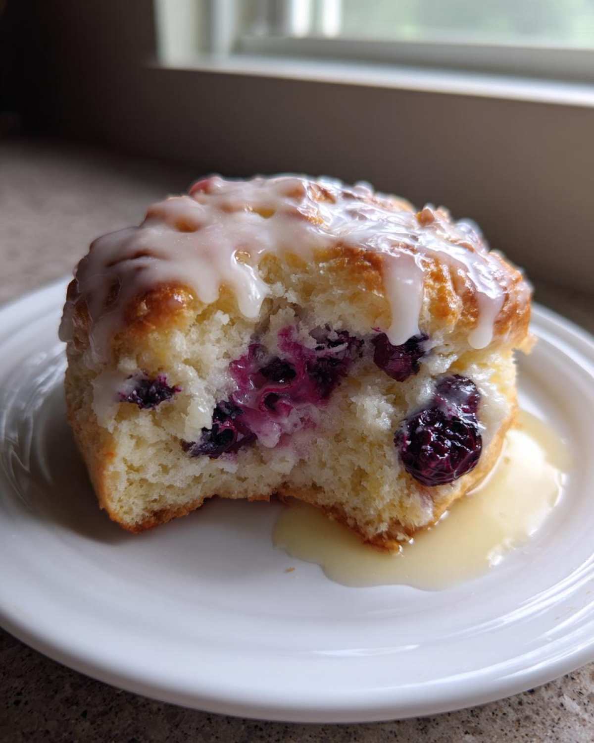 A close-up of a flaky Blueberry Lemon Scones half, drizzled with white glaze, sitting on a white plate.