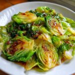 Close-up of vibrant green cabbage stir fry coated in a glossy garlic ginger sauce, served on a white plate.