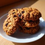 A stack of four chewy Gluten-Free Oatmeal Raisin Cookies resting on a white plate, bathed in sunlight.