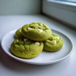 Four bright green Chewy Matcha White Chocolate Cookies piled on a white plate near a window.