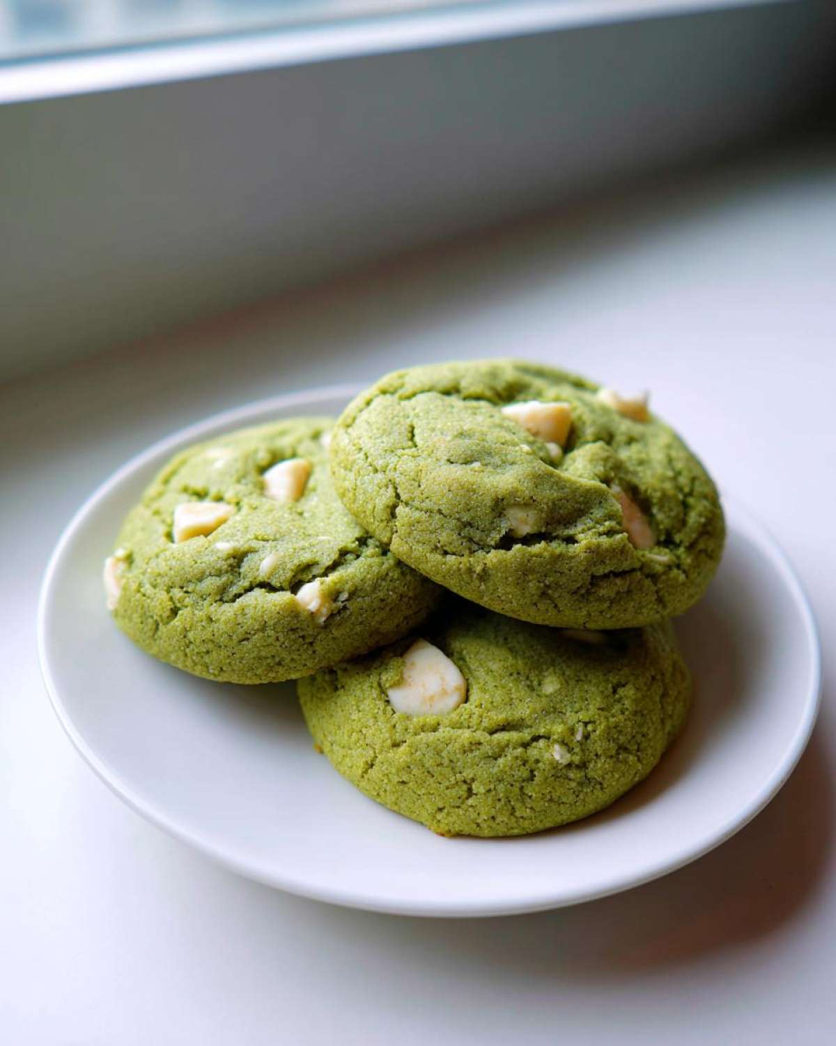 Three vibrant green Chewy Matcha White Chocolate Cookies studded with white chocolate chips, stacked on a small white plate.