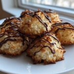 A close-up of several golden-brown Coconut Macaroons with Dark Chocolate Drizzle stacked on a white plate.