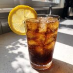 A tall glass filled with ice and dark, fizzy Cold Brew Lemonade, garnished with a bright lemon wheel.