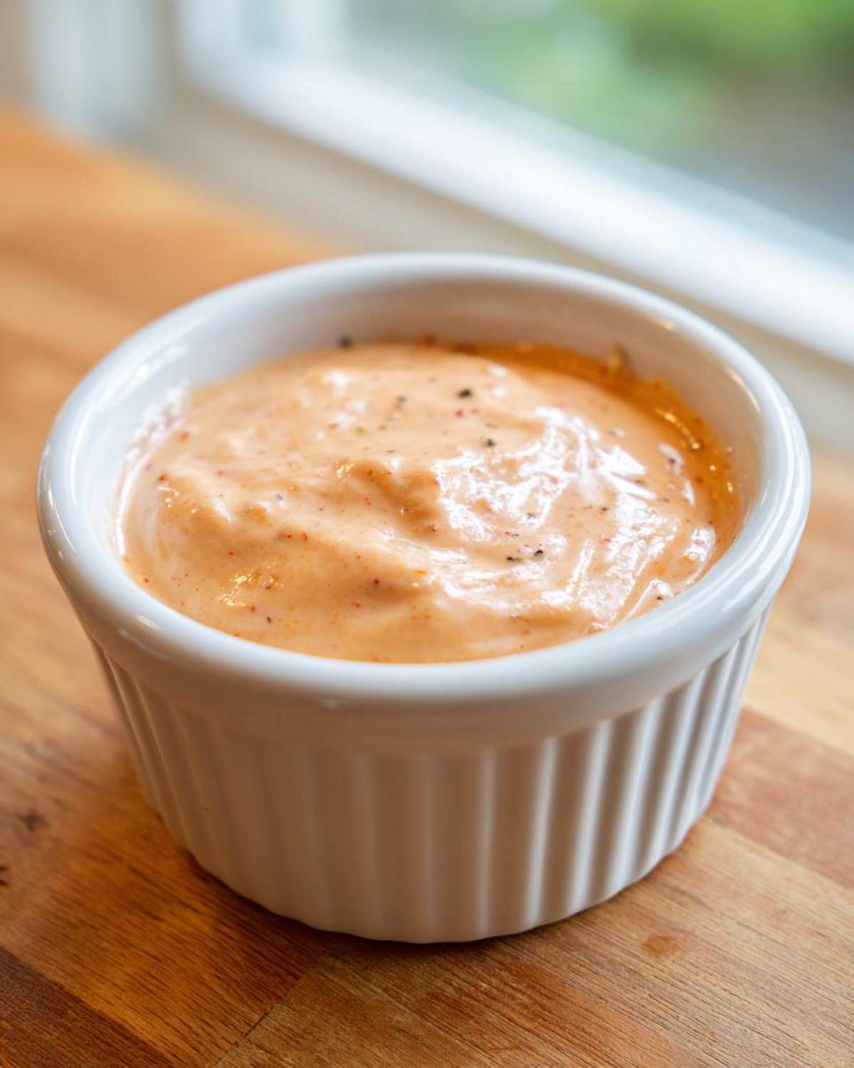 A close-up of creamy, orange-hued Copycat Raising Cane’s Sauce speckled with pepper in a small white ramekin.