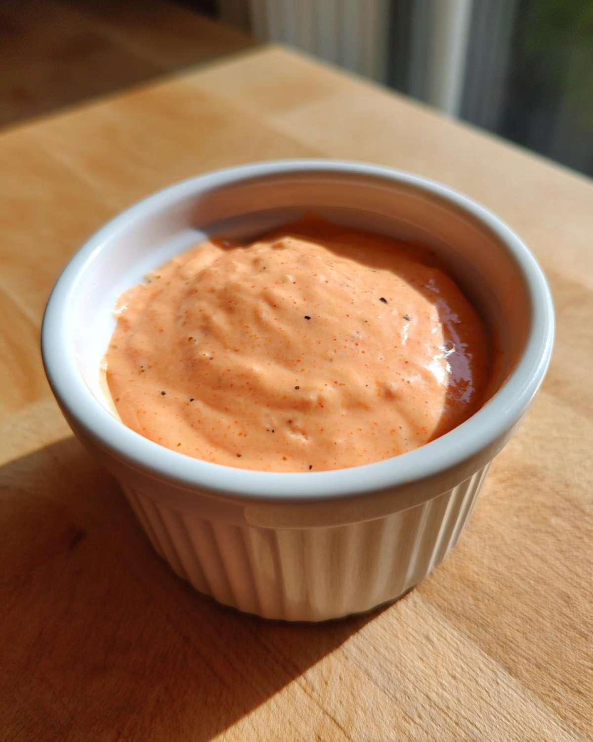 A small white ramekin filled with orange-hued Copycat Raising Cane’s Sauce, resting on a wooden cutting board.