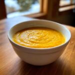 A close-up of a white bowl filled with thick, vibrant orange Creamy Coconut Lentil Soup on a wooden table.