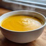 A close-up of a steaming white bowl filled with bright orange Creamy Coconut Lentil Soup, resting on a wooden surface.