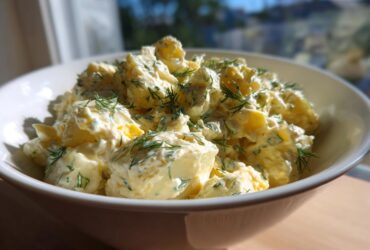 A close-up of a white bowl filled with Creamy Dill Potato Salad, generously topped with fresh dill fronds.