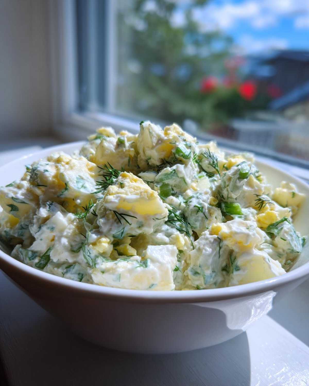 A white bowl filled with Creamy Dill Potato Salad featuring chunks of potato, egg, and fresh dill, set near a window.