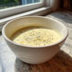 A white bowl filled with creamy yellow dressing, topped with shredded parmesan and dried herbs, resembling Copycat Olive Garden Salad Dressing.