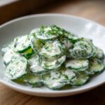 A white bowl filled with sliced cucumbers coated in a creamy dressing and topped with fresh dill, showcasing the Creamy German Cucumber Salad.