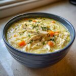 A close-up of a bowl of Creamy Lemon Chicken Orzo Soup, topped with shredded chicken, carrots, and parsley.
