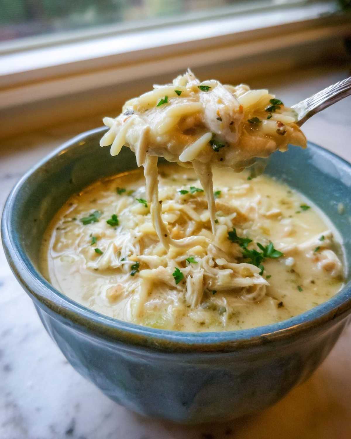 A spoonful of creamy lemon chicken orzo soup being lifted from a blue bowl, showing shredded chicken and orzo pasta.