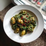 A white bowl filled with tender gnocchi coated in a rich, green creamy pesto sauce with spinach, topped with red chili flakes.