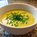 A close-up of a white bowl filled with bright yellow Creamy Potato Leek Soup, topped generously with fresh chopped chives.