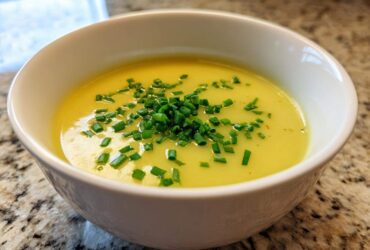 A close-up of a white bowl filled with bright yellow Creamy Potato Leek Soup, topped generously with fresh chopped chives.