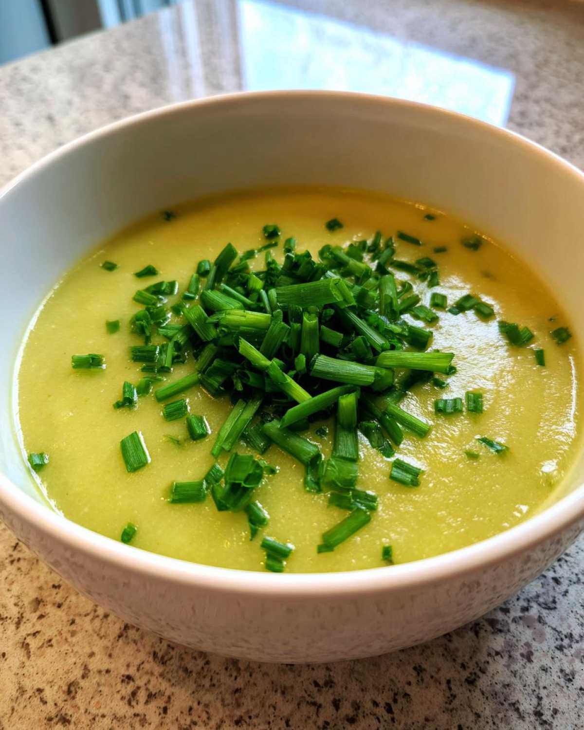 A close-up of a bowl of smooth, yellow-green Creamy Potato Leek Soup topped generously with fresh, chopped chives.