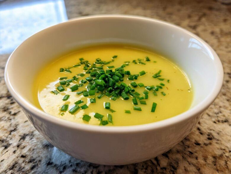 A close-up of a white bowl filled with bright yellow Creamy Potato Leek Soup, topped generously with fresh chopped chives.
