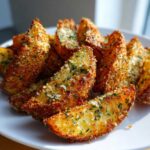 A close-up of golden brown Crispy Baked Garlic Parmesan Potato Wedges sprinkled with grated cheese and parsley on a white plate.
