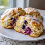Close-up of flaky Blueberry Lemon Scones, one split open showing juicy blueberries and topped with white lemon glaze.
