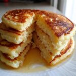 A stack of fluffy Gluten-Free Buttermilk Pancakes cut open to show the airy interior, drizzled with syrup.