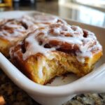 A close-up of a fluffy Gluten-Free Cinnamon Roll with a bite taken out, covered in white icing.