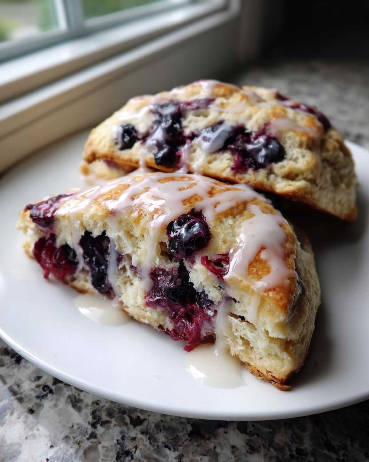 Two flaky Blueberry Lemon Scones drizzled with a thick white glaze, sitting on a white plate.