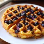 Close-up of a golden, crispy Gluten-Free Blueberry Waffles topped with juicy, cooked blueberries.
