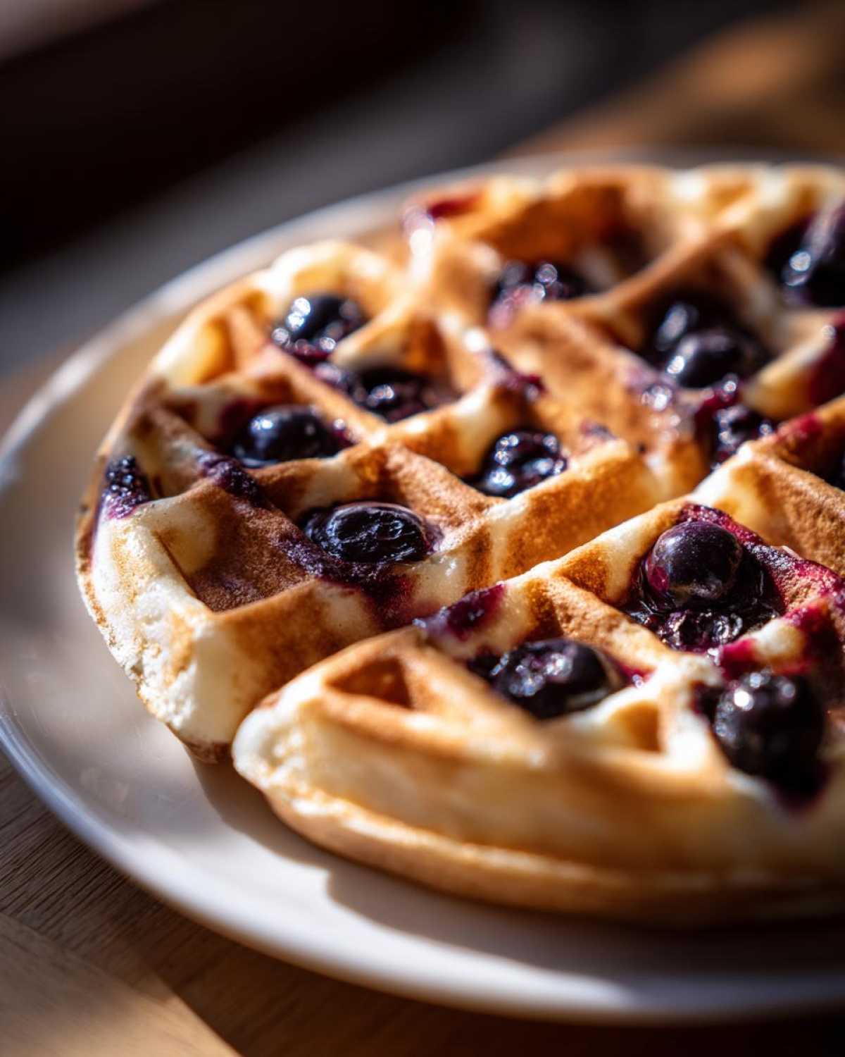 Close-up of golden Gluten-Free Blueberry Waffles studded with juicy, dark blueberries on a white plate.