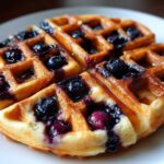Close-up of a golden, round Gluten-Free Blueberry Waffles topped with fresh blueberries and syrup on a white plate.