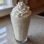 A tall glass filled with Homemade Vanilla Bean Frappuccino, speckled with vanilla seeds and topped high with whipped cream.