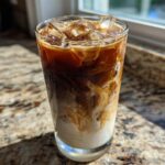 A tall glass of Iced Brown Sugar Oatmilk Latte showing rich layers of coffee, oatmilk, and ice cubes, glistening in sunlight.