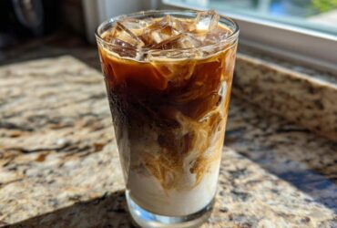 A tall glass of Iced Brown Sugar Oatmilk Latte showing rich layers of coffee, oatmilk, and ice cubes, glistening in sunlight.