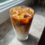 A tall glass showing the beautiful swirl of coffee mixing with almond milk in an Iced Honey Almond Milk Latte, topped with ice.