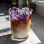 A glass containing an Iced Lavender Latte with swirling milk and espresso, topped with ice and purple lavender flowers.