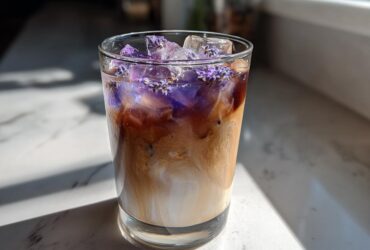 A glass containing an Iced Lavender Latte with swirling milk and espresso, topped with ice and purple lavender flowers.