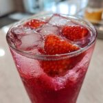 Close-up of a tall glass filled with vibrant red Iced Strawberry Lemon Tea, fresh strawberries, and ice cubes.