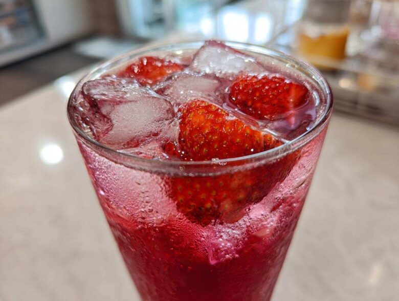 Close-up of a tall glass filled with vibrant red Iced Strawberry Lemon Tea, fresh strawberries, and ice cubes.