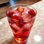 Close-up of a tall glass filled with vibrant red Iced Strawberry Lemon Tea, ice cubes, and fresh strawberry slices.
